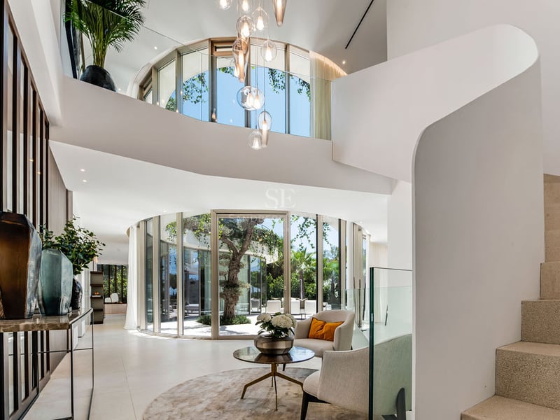 Entrée de luxe avec parois de verre courbes, patio central, escalier blanc sculptural et sol en pierre naturelle.