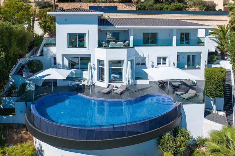 Vue aérienne d'une villa blanche moderne avec une grande piscine à débordement bleue et des terrasses meublées.