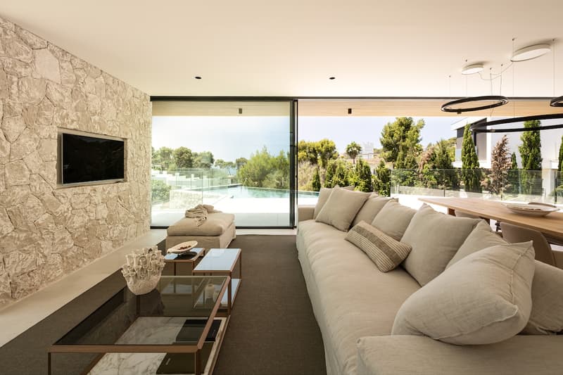 Salón de lujo moderno con pared de piedra natural Salón moderno con pared de piedra, sofá beige y puertas de cristal con vistas a la piscina y jardín.