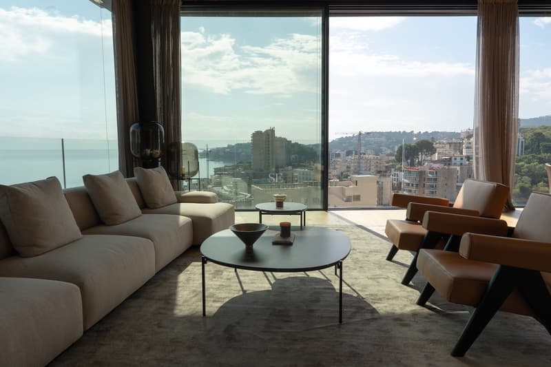 Modernes Wohnzimmer mit bodentiefen Fenstern, Blick auf die Küstenstadt, beiges Sofa und Lederstühle.