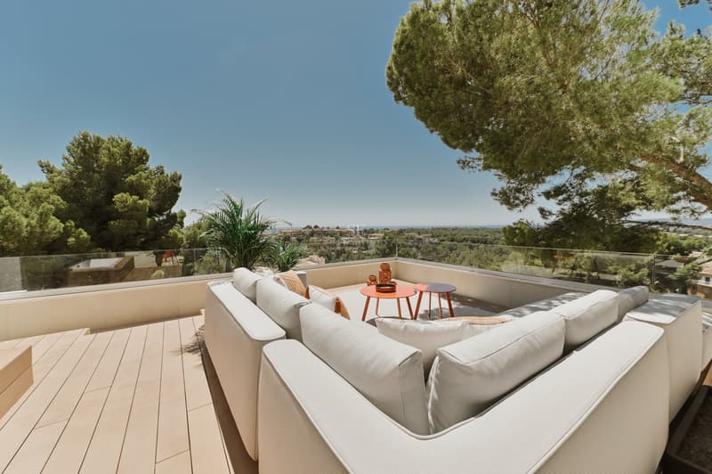 Sofá modular blanco en terraza de madera con barandillas de cristal y vistas al paisaje mediterráneo y al mar.