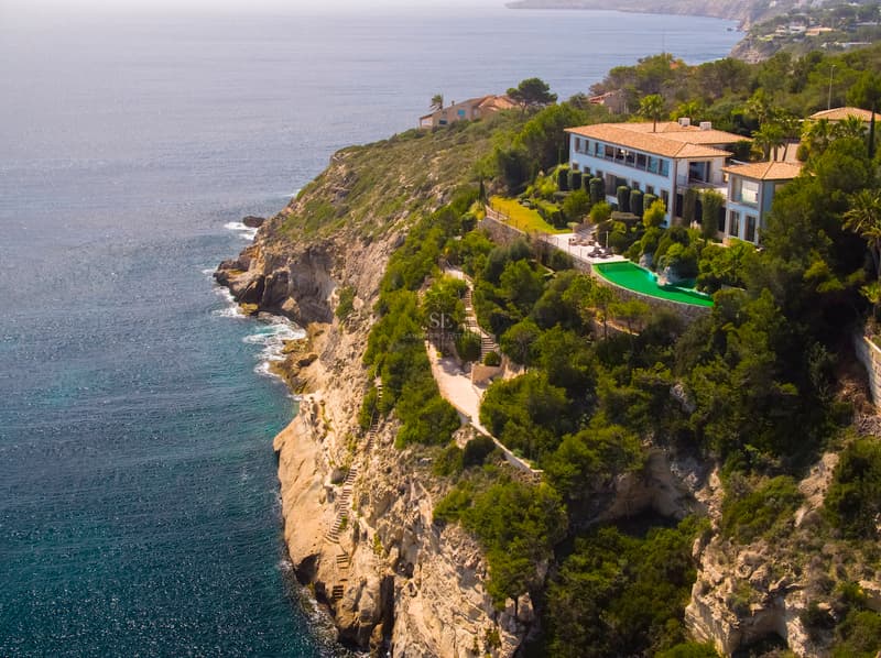 Exklusiv villa på klippavsats med havsutsikt Flygbild av en lyxvilla på en klippa vid havet med en rundad grön pool och panoramautsikt.