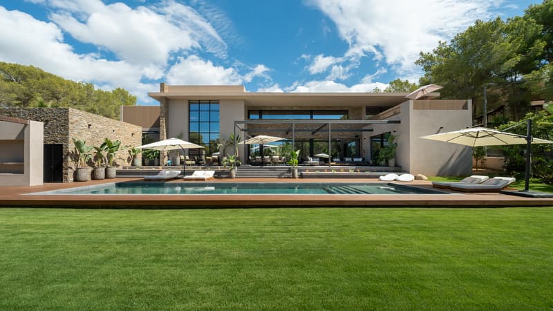 Villa contemporánea con piscina alargada, plataforma de madera, césped verde y fachada de piedra bajo un cielo azul.
