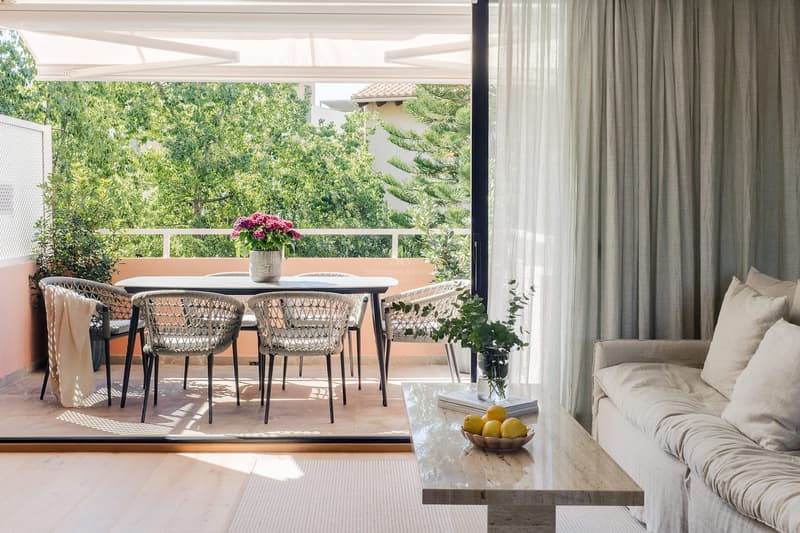 Elegant Terrass och Vardagsrum med Trädgårdsutsikt Utsikt från ett ljust vardagsrum mot en möblerad terrass med matbord, omgiven av grönskande träd.