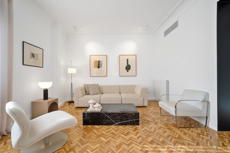 Salón luminoso con paredes blancas, suelo de parqué en espiga, sofá beige y una llamativa mesa de mármol negro.