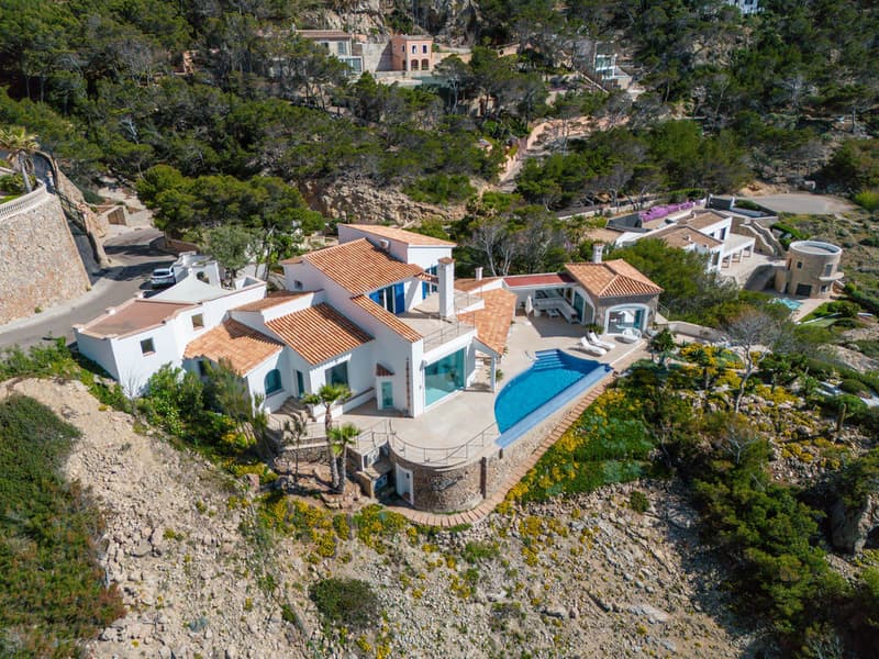 Aerial view of a white multi-level villa with terracotta roofs, infinity pool, and stone walls on a lush hillside.