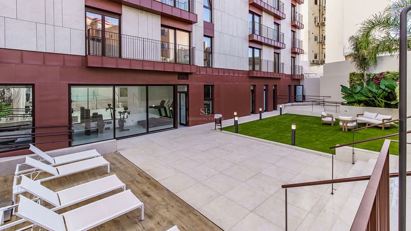 Modern innergård med solstolar, gräsmatta och inglasat gym vid foten av en samtida byggnad.