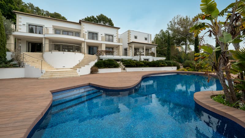 Außenansicht einer weißen Luxusvilla mit großem geschwungenem Pool und Holzdeck, umgeben von mediterraner Natur.