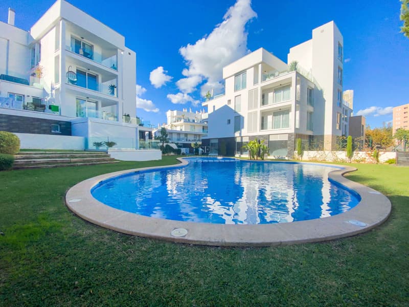 Residencial Moderno con Piscina y Jardines Piscina azul curva rodeada de césped y edificios blancos modernos bajo un cielo despejado con nubes.