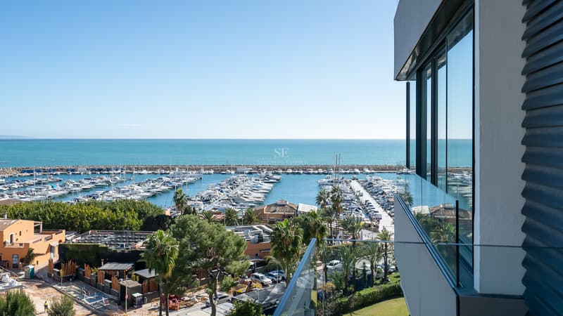 Vistas Panorámicas al Puerto Deportivo y al Mar Vista elevada desde un balcón moderno hacia un puerto deportivo lleno de yates y el mar Mediterráneo bajo un cielo despejado.