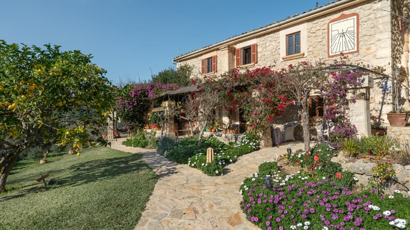 Fassade einer Natursteinfinca mit Fensterläden aus Holz, einem Steinweg und blühenden Bougainvillea-Pflanzen.