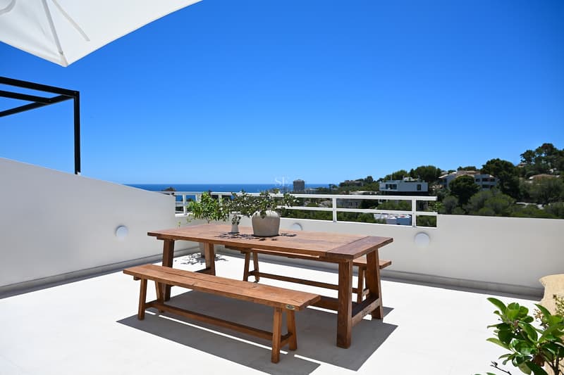 Mesa de comedor de madera en terraza blanca con vistas al mar Mediterráneo bajo un cielo azul despejado.