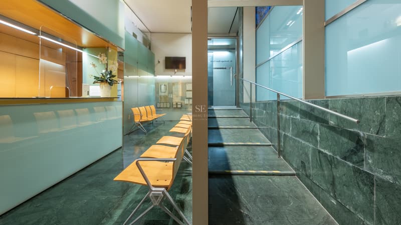 Zona de recepción con suelo de mármol verde, sillas de madera, paneles de cristal esmerilado y escalera metálica.