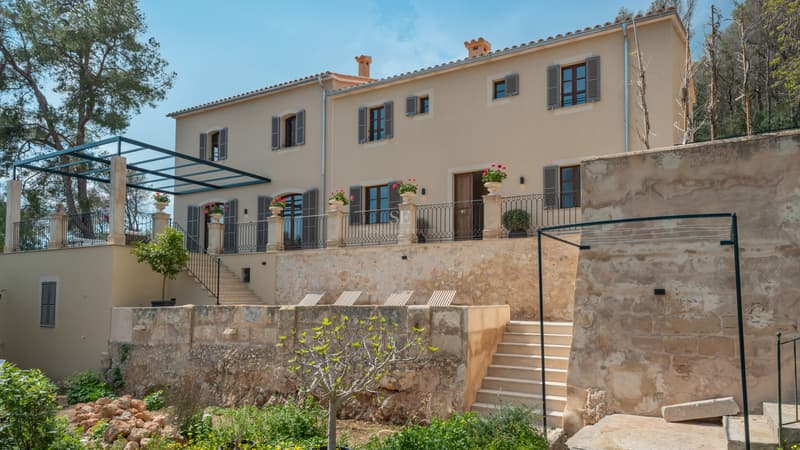 Elegante villa mediterránea beige de dos plantas con persianas gris azulado, muros de terraza de piedra y barandillas.