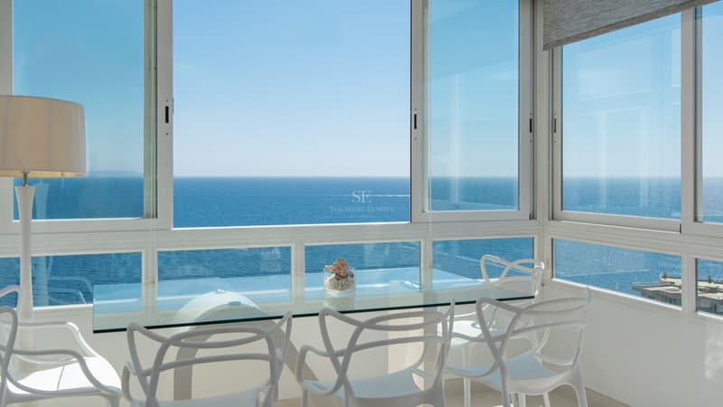 Modern dining area with white designer chairs and glass table overlooking the calm blue Mediterranean sea.