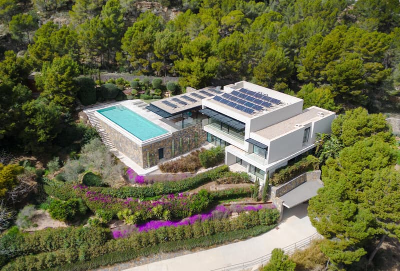 Vue aérienne d'une villa moderne blanche avec piscine à débordement, panneaux solaires et forêt de pins.