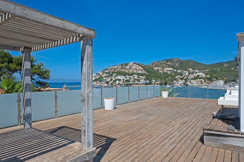 Terraza de madera en azotea con pérgola y vistas panorámicas al mar, puerto y villas en la montaña bajo un cielo despejado.
