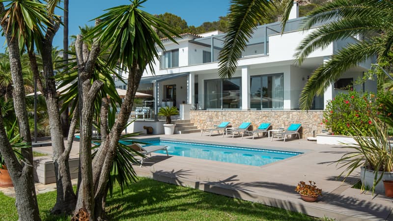 Villa Contemporánea con Piscina y Jardín Mediterráneo Villa blanca moderna con piscina rectangular, tumbonas y palmeras bajo un cielo despejado.