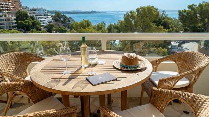 Terraza elevada con mesa de madera, sillas de mimbre y vistas al mar azul entre pinos mediterráneos.