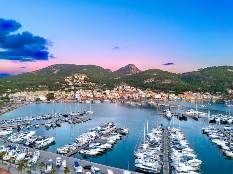 Vista aérea de un puerto deportivo de lujo con yates blancos bajo un cielo rosa y azul junto a montañas verdes.