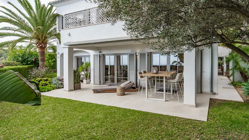 Elegant Covered Terrace and Mediterranean Garden White Mediterranean villa with a covered patio, high dining table, sun lounger, green lawn, and palm trees.