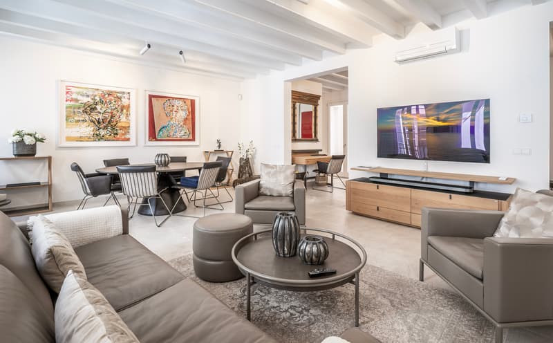 Amplio salón moderno con sofás de cuero gris, vigas de madera, televisor grande y zona de comedor con arte.
