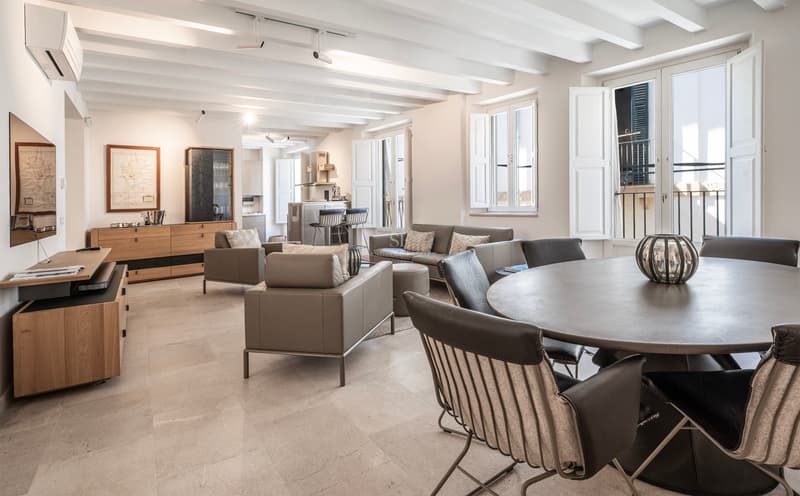 Amplio salón con techos de vigas blancas, muebles de cuero gris y una mesa de comedor redonda en primer plano.