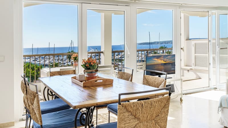 Comedor soleado con mesa de madera y sillas de mimbre con vistas a un puerto lleno de yates y al océano azul.