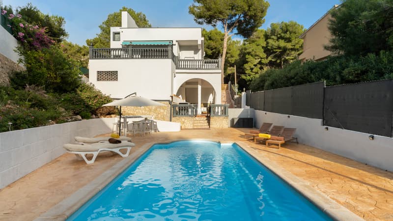 Blå pool omgiven av en stenterrass med solstolar och en vit villa i bakgrunden under en klarblå himmel.