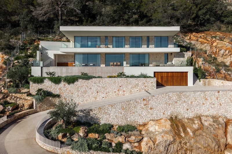 Villa blanca moderna en una colina rocosa con muros de piedra seca, grandes ventanales y un camino de entrada curvo.