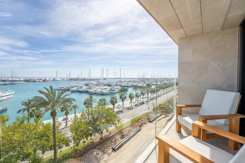 Terraza moderna con sillas de madera y barandilla de cristal con vistas a un puerto lleno de yates y palmeras.