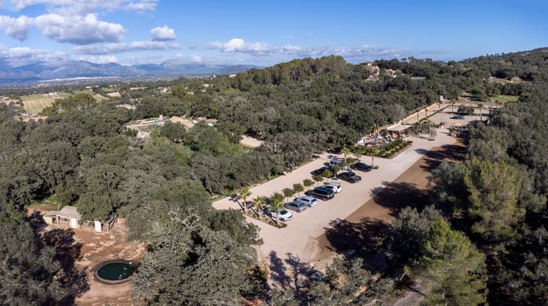 High-angle drone shot of a large Mediterranean estate with ample parking, surrounded by dense green forest and mountains.