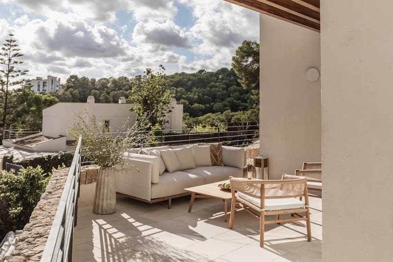 Terraza exterior con sofá moderno, mesa de madera y sillas de cuerda con vistas a un valle verde.