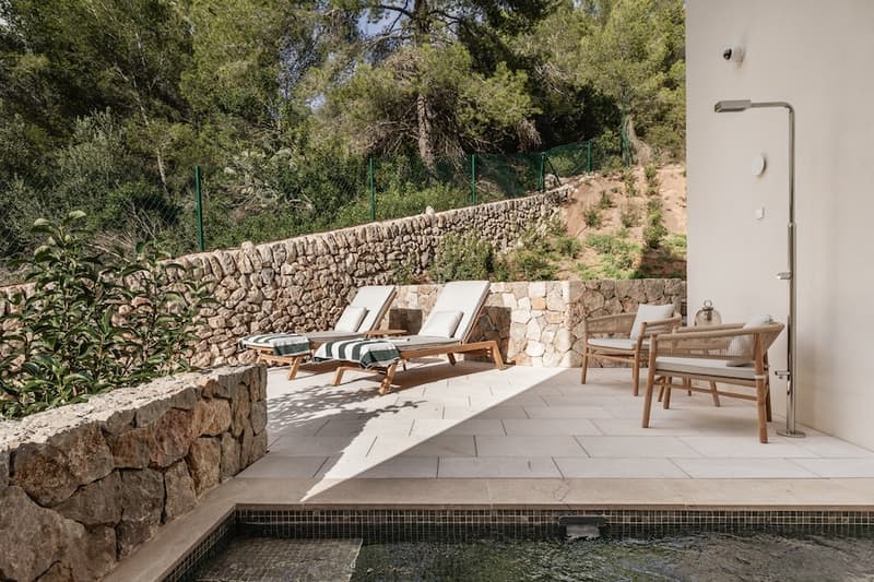 Terraza moderna de piedra con tumbonas, una ducha exterior metálica y vistas a un bosque frondoso.