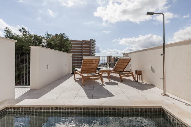 Sonnige Terrasse mit kleinem Pool, zwei Sonnenliegen aus Holz und Außendusche unter blauem Himmel.