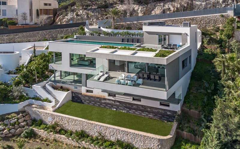 Flygfoto över en vit modern flervåningsvilla med en infinitypool på taket och glasterrasser.