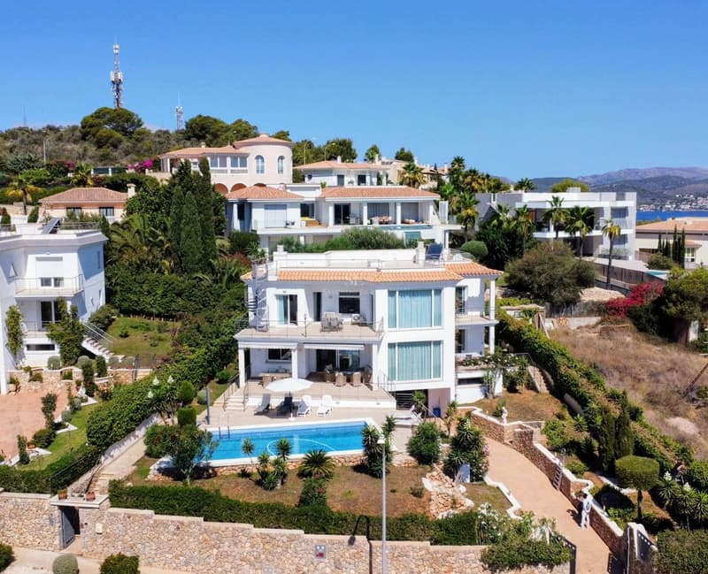Aerial shot of a white multi-level villa featuring a rectangular turquoise pool, stone walls, and manicured gardens.