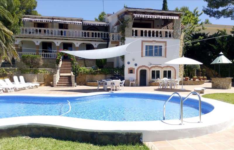 Gran piscina curvada frente a una villa blanca de estilo mediterráneo con terrazas de terracota y muros de piedra.