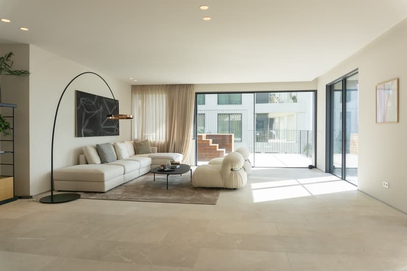 Ljust minimalistiskt vardagsrum med beige stengolv, off-white hörnsoffa och stora skjutdörrar i glas.