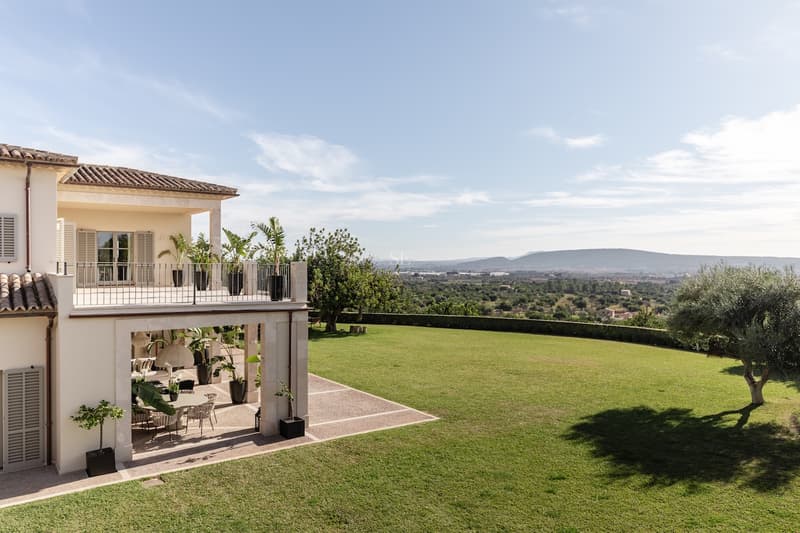 Villa Méditerranéenne avec Grand Jardin et Vue Villa crème avec vaste pelouse, terrasse ornée de palmiers et vue panoramique sur les montagnes par temps clair.