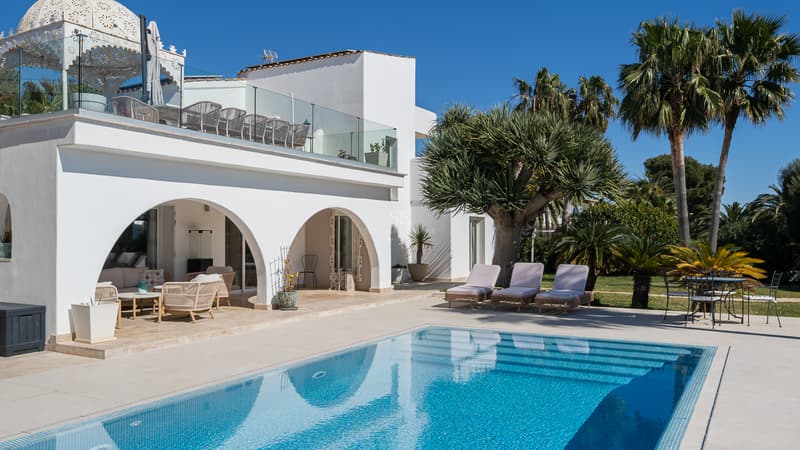 Villa méditerranéenne de luxe avec piscine privée Villa blanche à arches surplombant une piscine turquoise avec chaises longues et palmiers sous un ciel bleu.