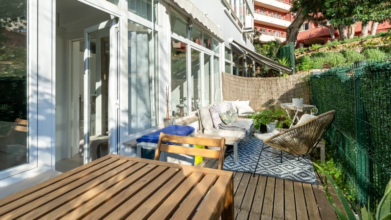 Terrasse privée en bois avec mobilier de jardin, tapis géométrique et baies vitrées sous une lumière naturelle vive.