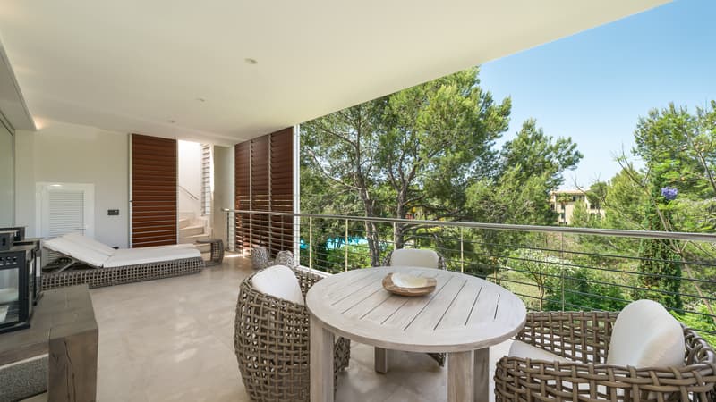 Terrasse couverte avec table en bois, fauteuils tressés et chaises longues donnant sur des pins.