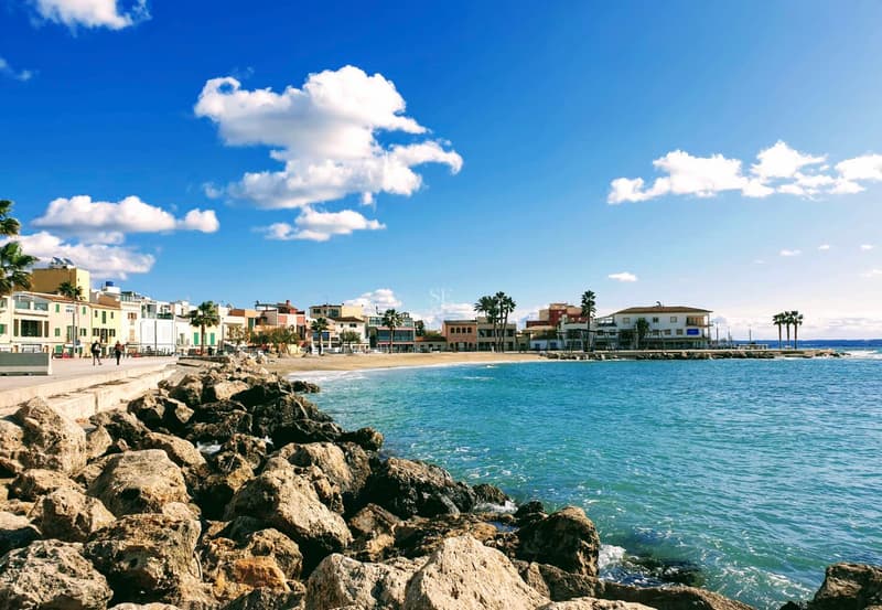 Vues Panoramiques en Bord de Mer, Emplacement Privilégié Vue depuis un rivage rocheux vers une plage de sable et des bâtiments côtiers sous un ciel bleu vif.