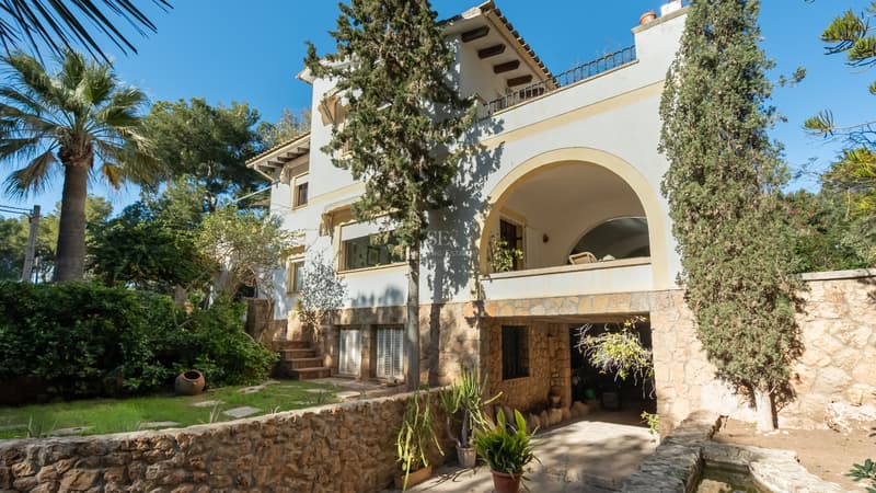 Villa méditerranéenne de trois étages avec balcon arqué, base en pierre naturelle et jardin mature sous un ciel bleu.
