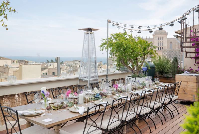 Una mesa de comedor elegantemente decorada en una terraza de madera con vistas a una ciudad histórica y al mar.