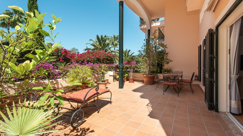 Terrasse en terre cuite avec chaise longue et coin repas, entourée de bougainvilliers et de palmiers sous un ciel bleu.