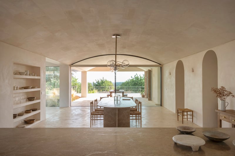 A minimalist dining room featuring a vaulted stone ceiling, oval marble table, and large glass doors to a sunlit terrace.
