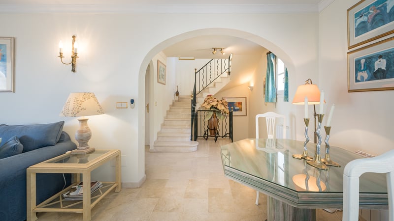 Vue depuis un salon avec table en verre et canapé bleu vers une arche et un escalier en marbre.