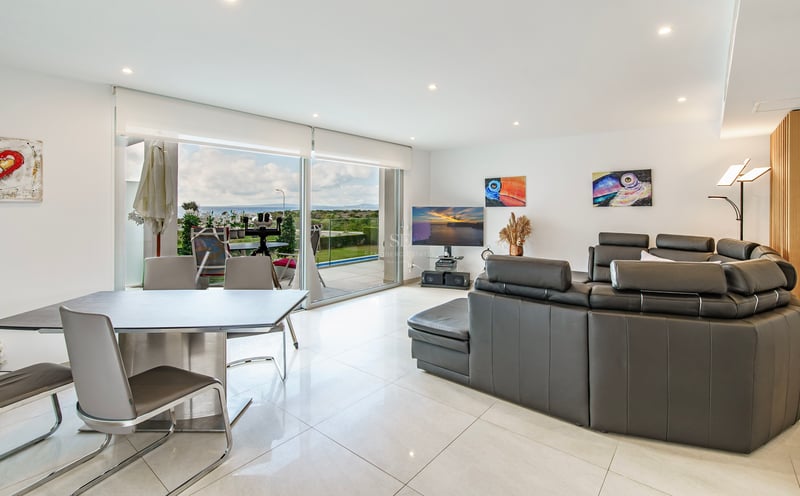 Salon moderne avec canapé en cuir gris foncé, coin repas et grandes baies vitrées ouvrant sur une vue mer.